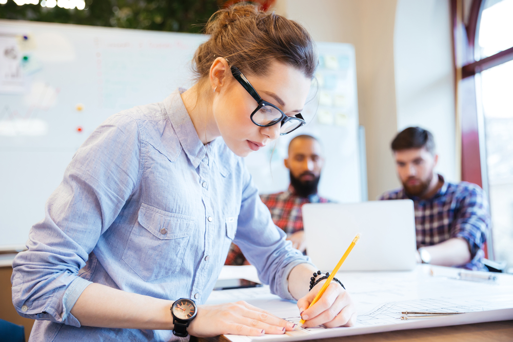 Woman engineer working on blueprint in office with colleagues on background